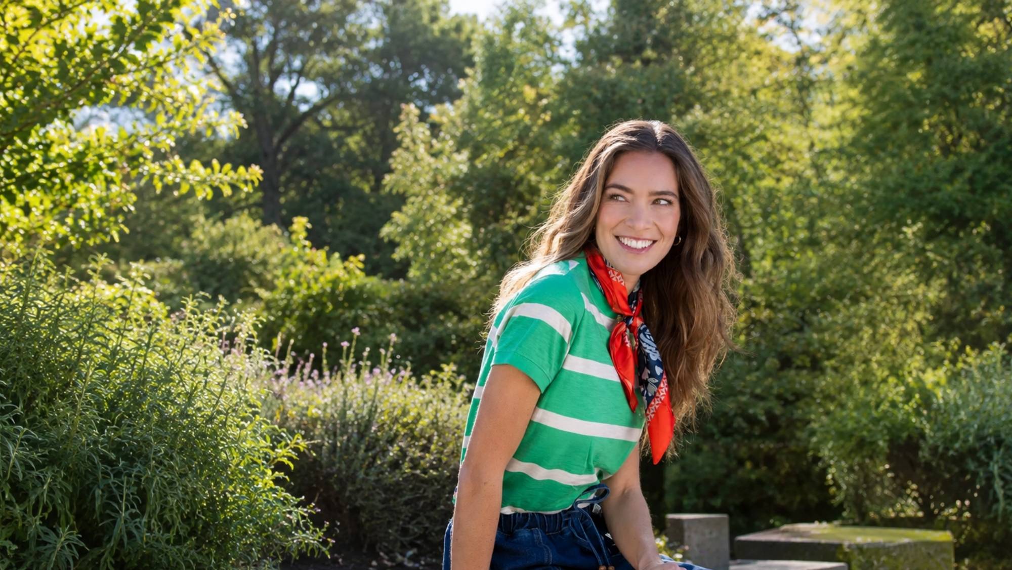 Smiling woman wearing a green striped top, red patterned scarf and blue jeans in a summer garden