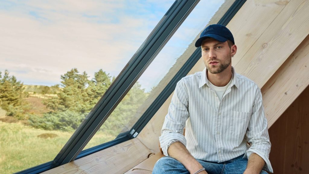 Man wearing a striped shirt, jeans and a navy cap beside a large window
